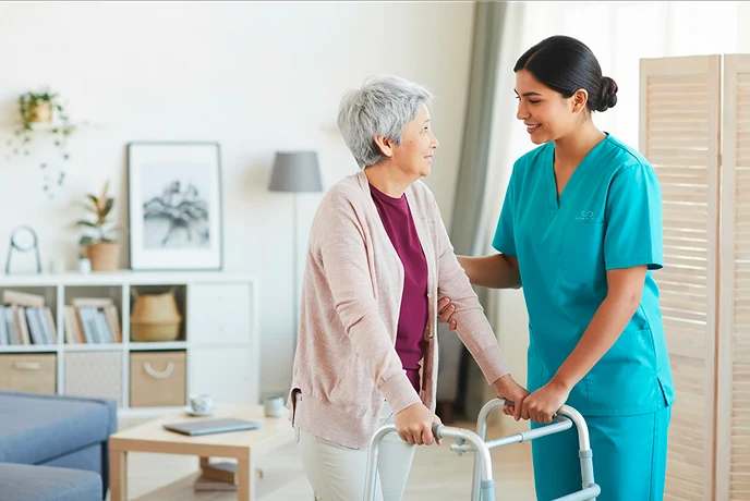 A smiling nurse in turquoise scrubs assists an elderly woman with a walker in a cozy, light-filled living room. The atmosphere is warm and supportive.