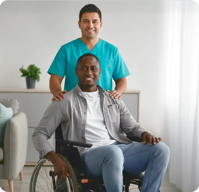A man in a wheelchair smiles warmly, accompanied by a caregiver in teal scrubs who stands behind, signifying support and care in a cozy room setting.
