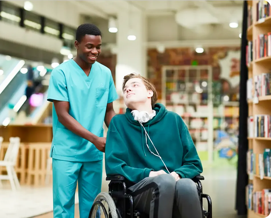 A caregiver in blue scrubs smiles while pushing a young man in a wheelchair through a bright library. Both appear happy and engaged in conversation.