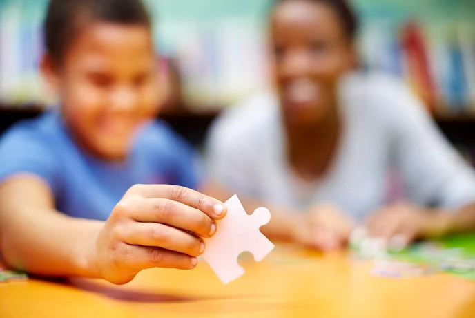 Child holding a puzzle piece while a teacher smiles in the background in a classroom.