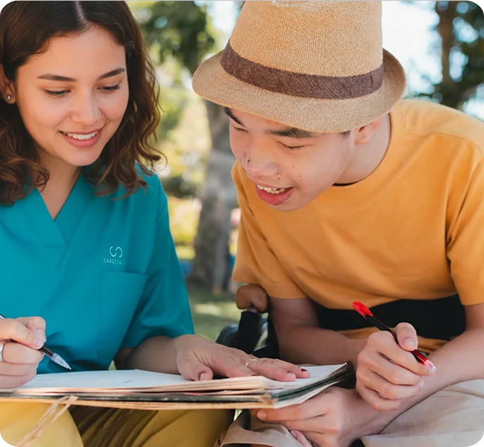 A young woman in a blue uniform and a young man in a hat are joyfully drawing together outdoors. They are both smiling, conveying a sense of collaboration and happiness.