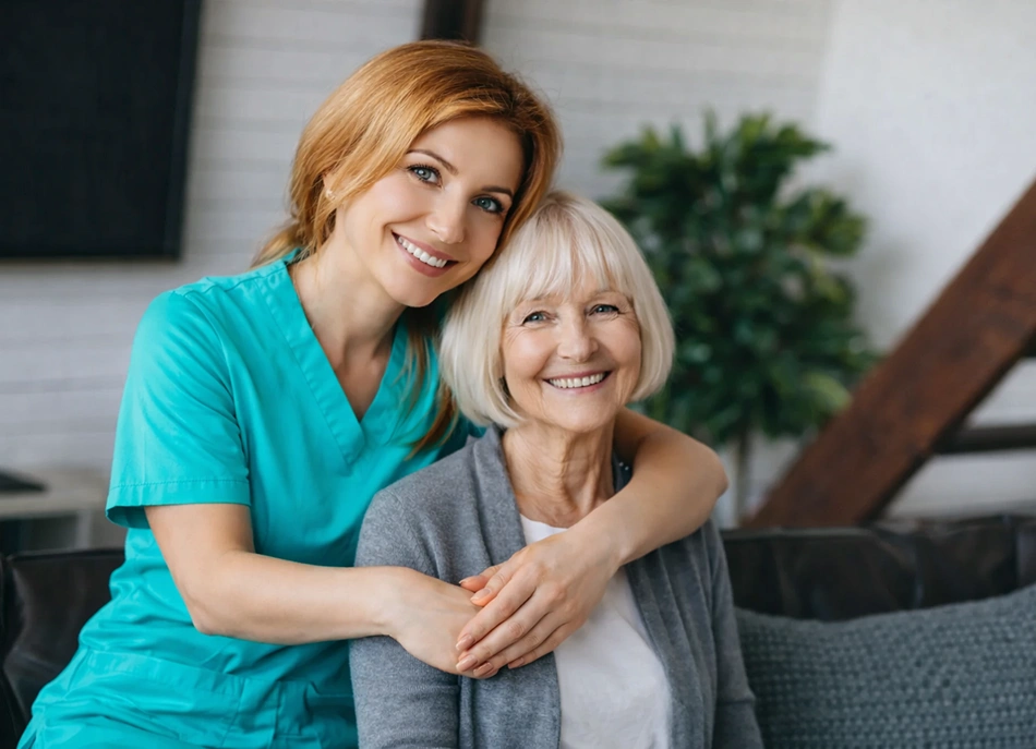 A smiling healthcare worker in teal scrubs embraces an elderly woman, both appearing joyful and content. They're seated in a cozy, well-lit room.