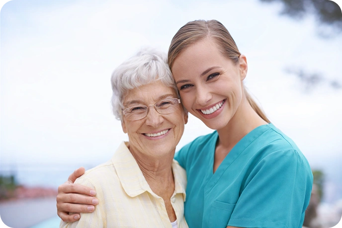 A smiling elderly woman with white hair stands arm-in-arm with a cheerful young woman in scrubs, conveying warmth and companionship outdoors.