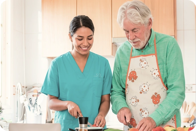 A caregiver in teal scrubs and an elderly man in a chicken-patterned apron smile while chopping vegetables in a bright kitchen. Warm and cheerful mood.