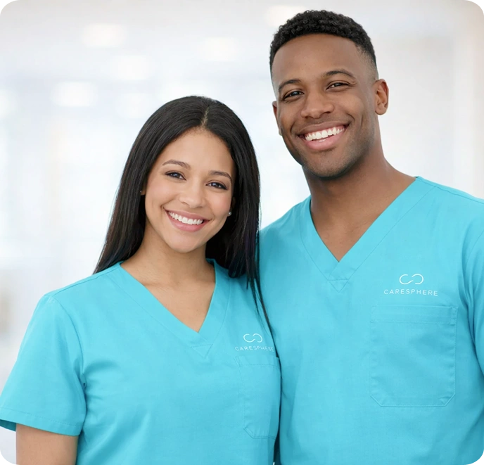 A smiling man and woman wearing turquoise scrubs stand closely together. The bright, blurred background suggests a professional healthcare setting.