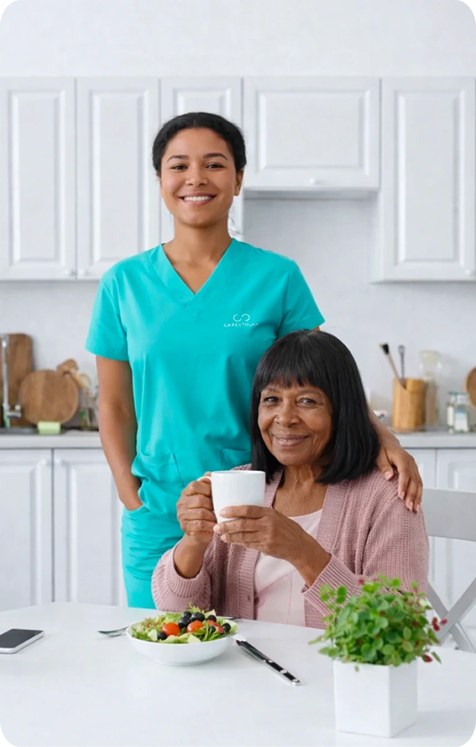 A smiling healthcare worker in teal scrubs stands beside an elderly woman holding a cup, seated at a kitchen table with a salad and plant, conveying warmth.