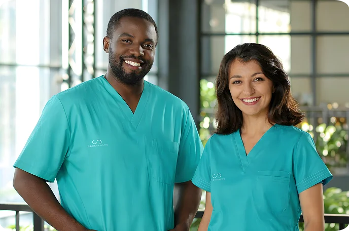 Two smiling healthcare professionals wearing turquoise scrubs stand in a well-lit hospital corridor, conveying a sense of friendliness and competence.