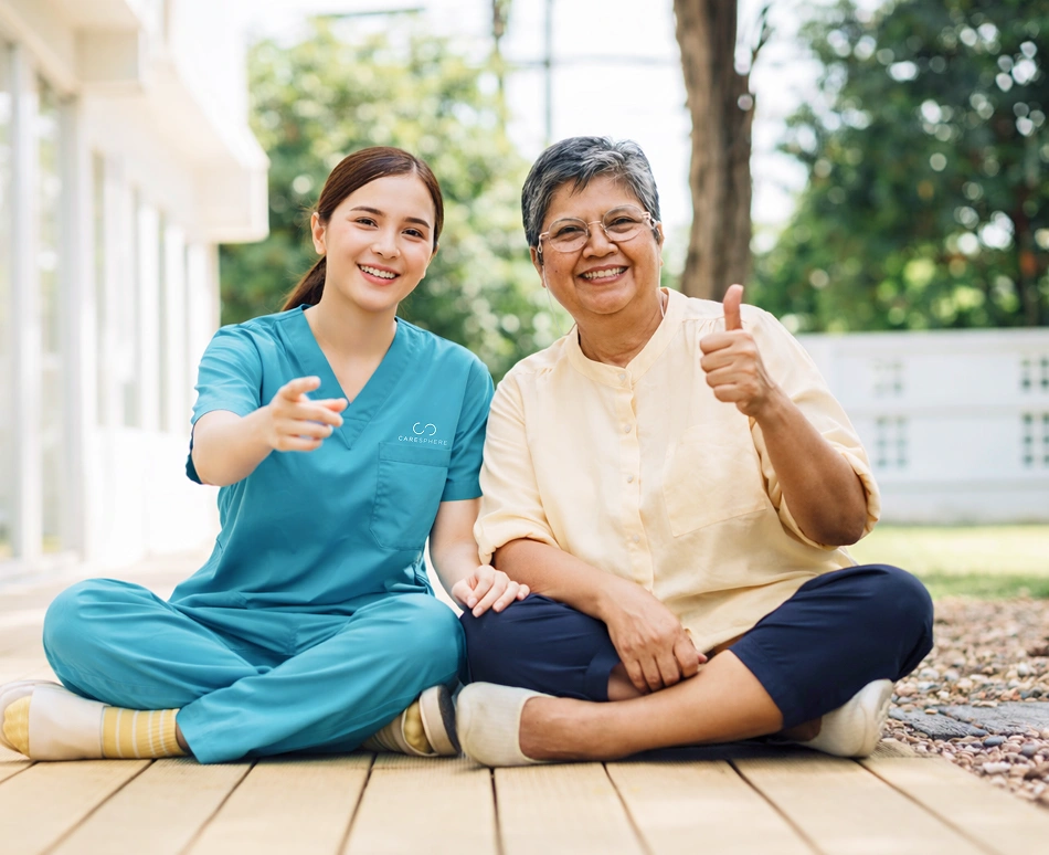 Caregiver sitting outdoors with a smiling senior woman giving a thumbs-up.