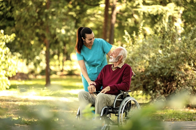 Female caregiver walking with an elderly man in a wheelchair in a park, sharing a warm moment outdoors