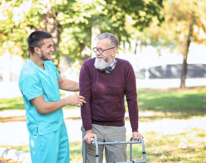 Caregiver assisting an elderly man using a walker during a walk in the park