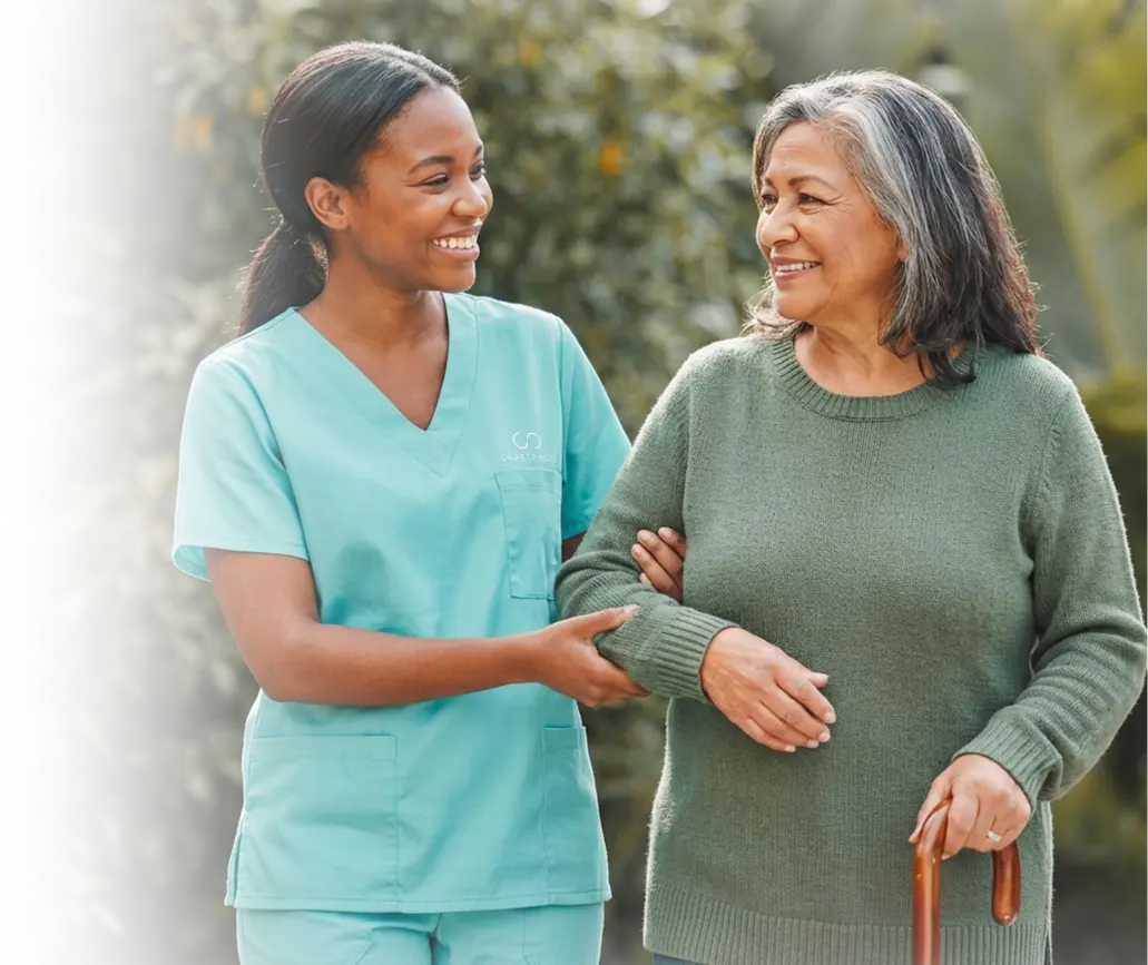 Caregiver in teal scrubs walking arm in arm with an elderly woman holding a cane outdoors
