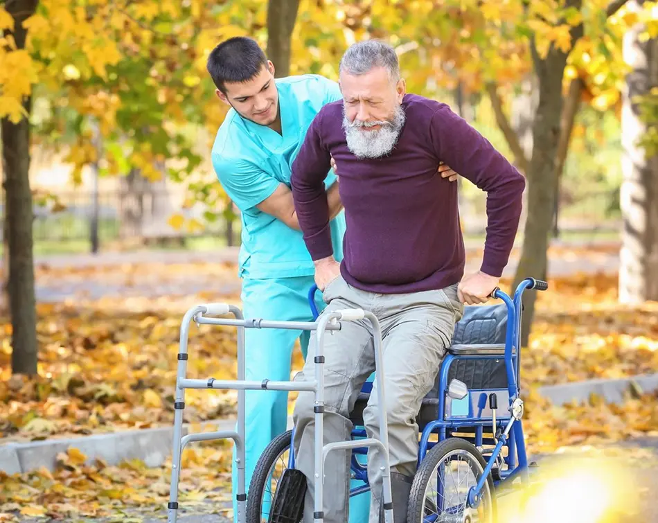 Caregiver in teal scrubs helping an elderly man rise from a wheelchair with a walker in an autumn park