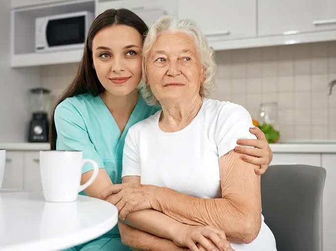 Caregiver in teal scrubs embracing an elderly woman at a kitchen table with a mug
