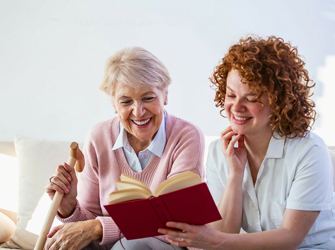 An elderly woman and a caregiver sitting together, smiling while reading a book, suggesting companionship and supportive care.