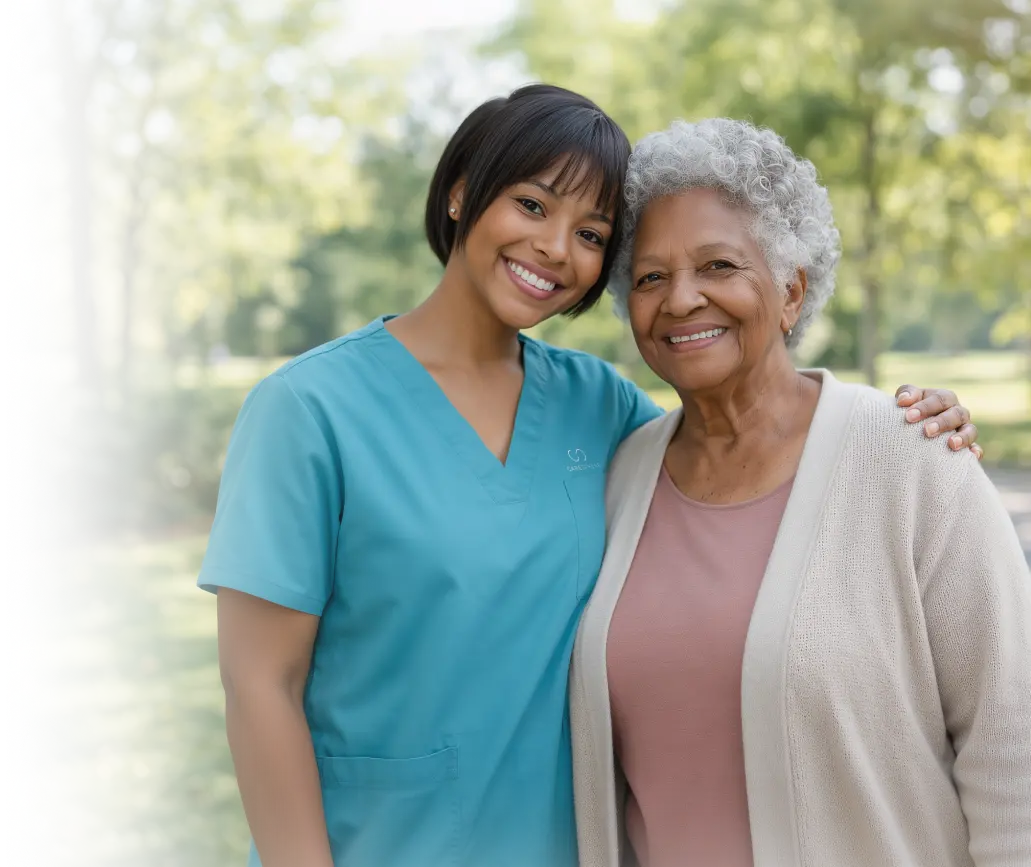 Caregiver in teal scrubs with her arm around a smiling elderly woman outdoors in a green park setting
