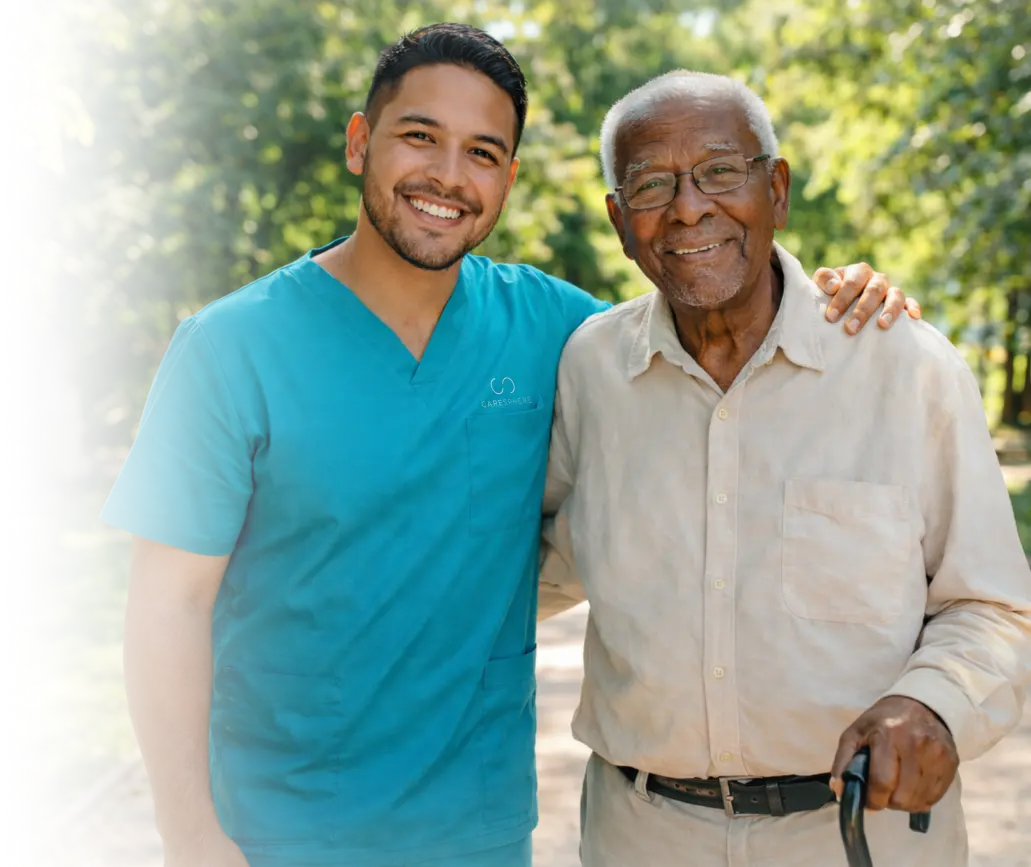 Smiling caregiver in teal scrubs with his arm around an elderly man with glasses and a cane outdoors