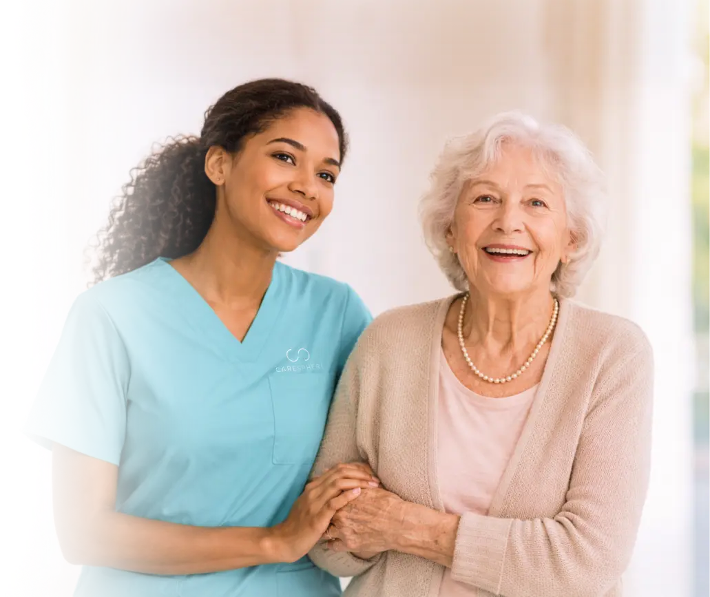Smiling caregiver in teal scrubs holding hands with a laughing elderly woman wearing a pearl necklace