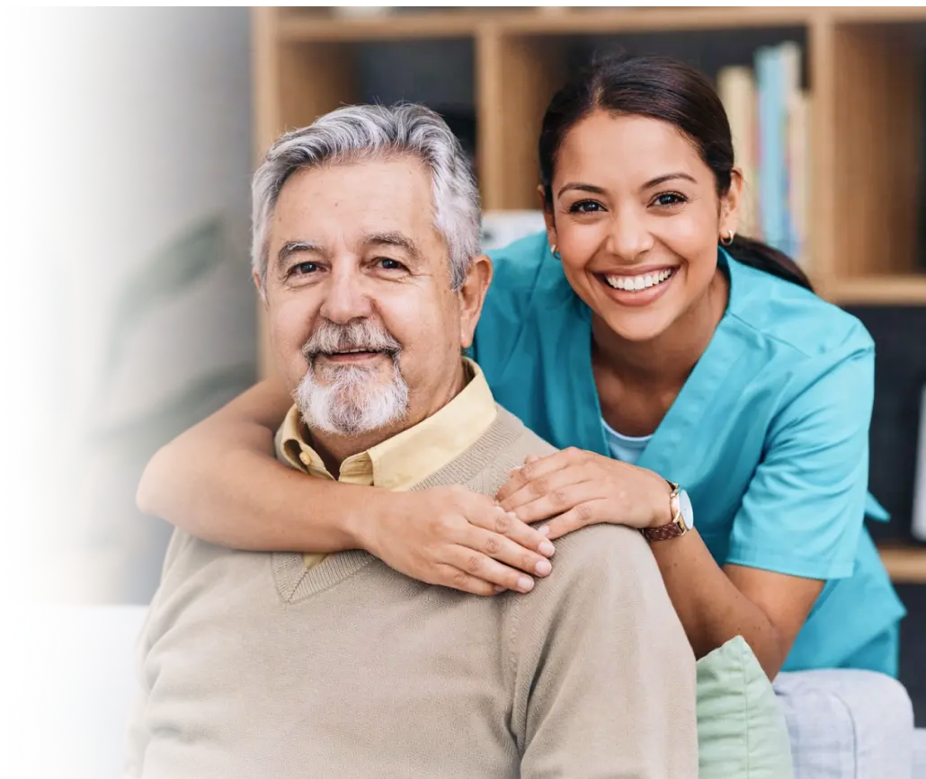 Smiling caregiver in teal scrubs leaning over the shoulder of a cheerful elderly man indoors