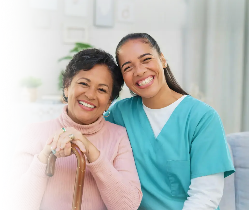 Laughing caregiver in teal scrubs cheek to cheek with a smiling elderly woman holding a cane