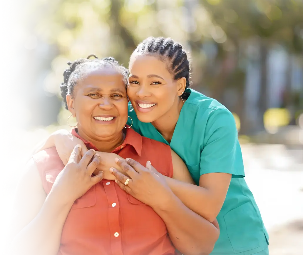 Caregiver in teal scrubs embracing a smiling elderly woman from behind outdoors in warm sunlight