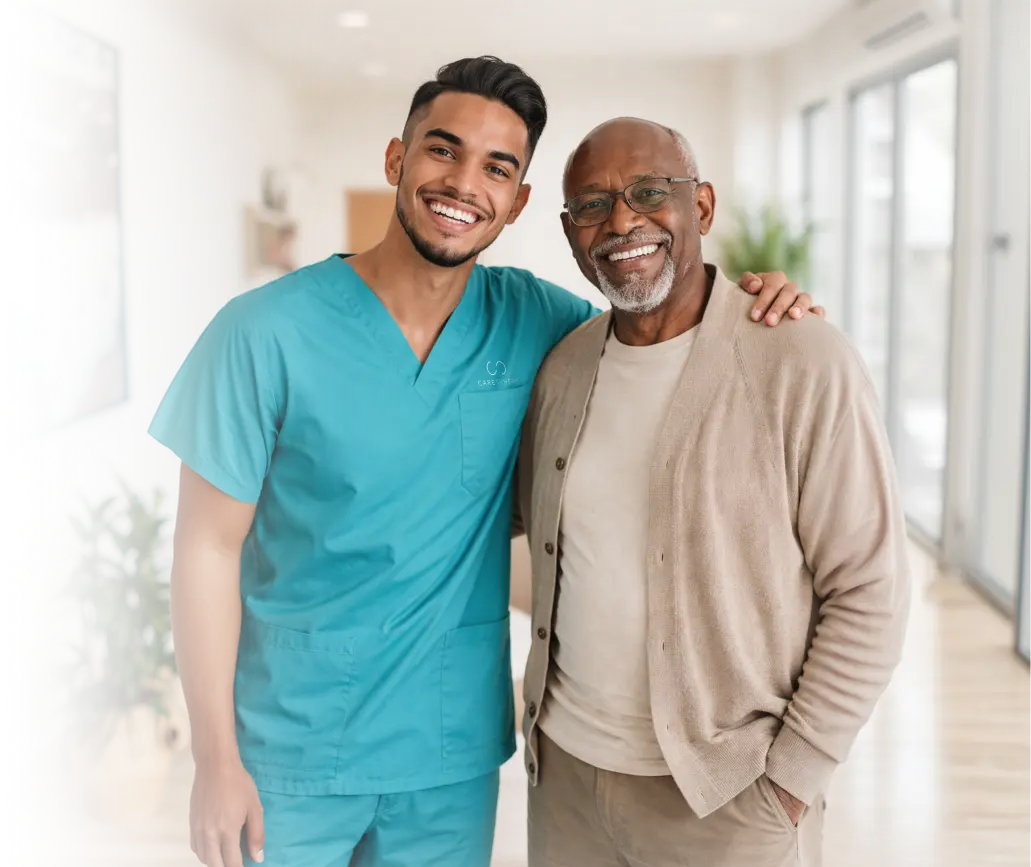 Smiling caregiver in teal scrubs standing with his arm around a cheerful elderly man with glasses in a bright hallway