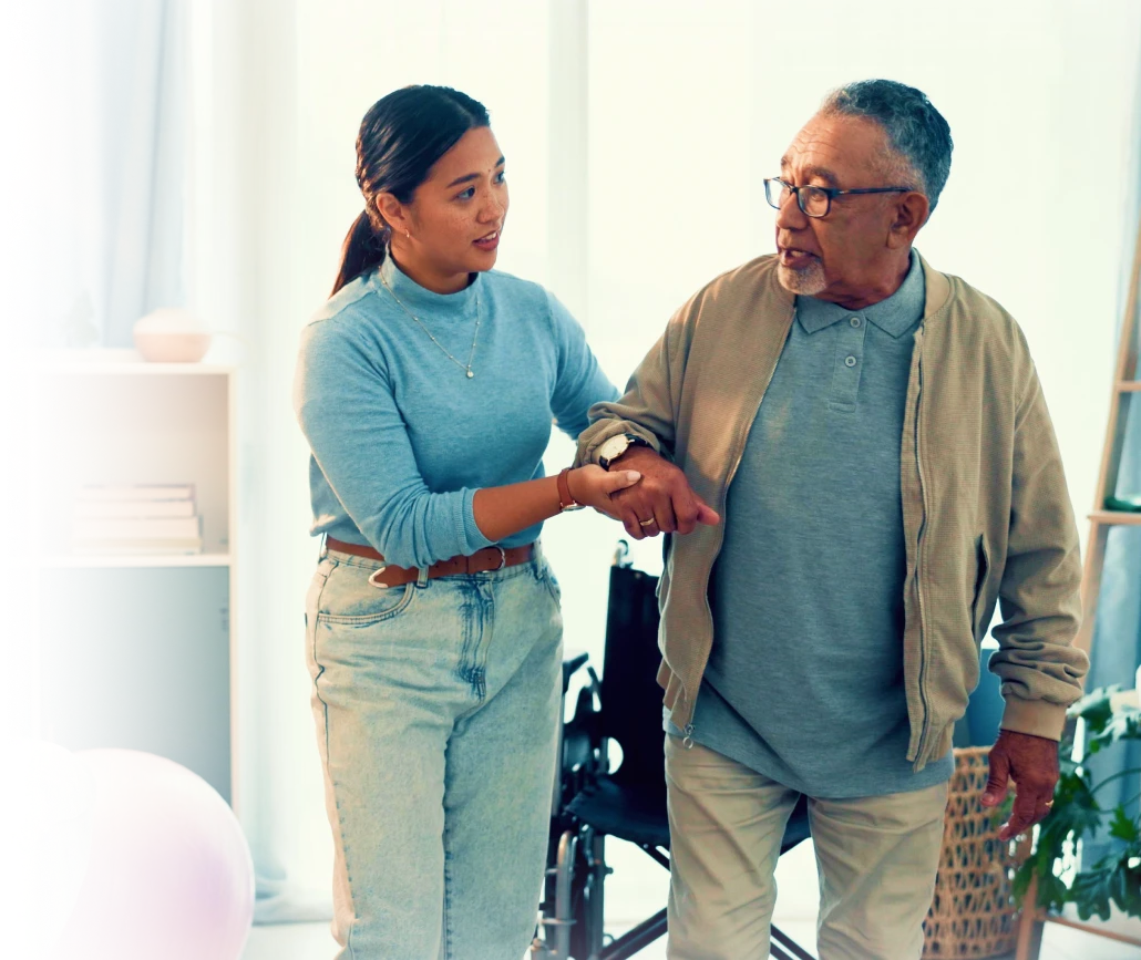 Caregiver assisting an elderly man with walking at home, with a wheelchair visible in the background