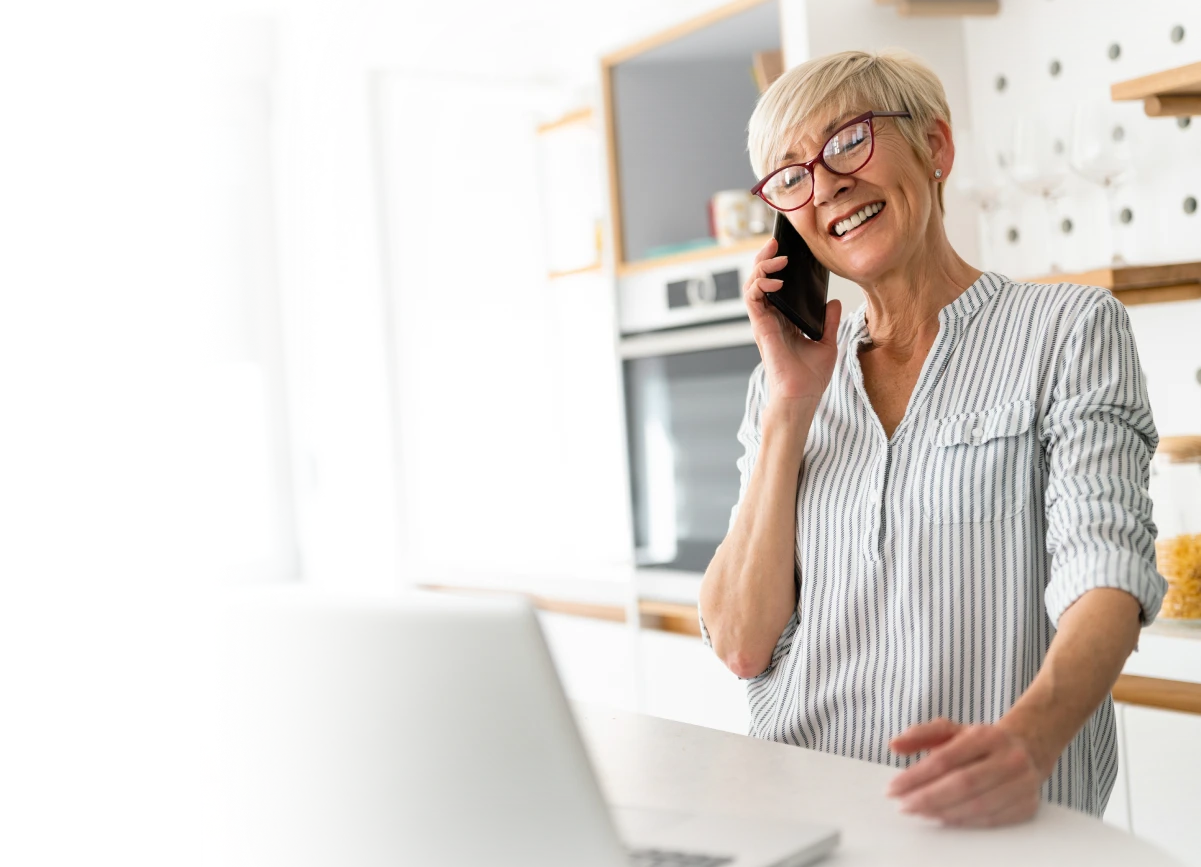 Smiling elderly woman with glasses talking on a phone while standing at a kitchen counter with a laptop