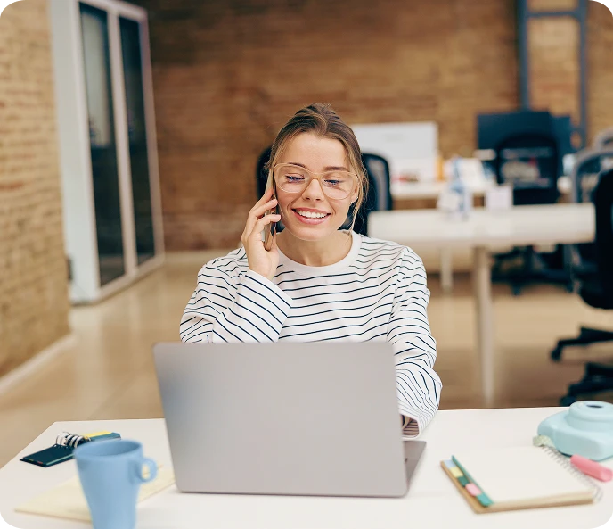 Smiling young woman with glasses talking on a phone while working on a laptop at an office desk